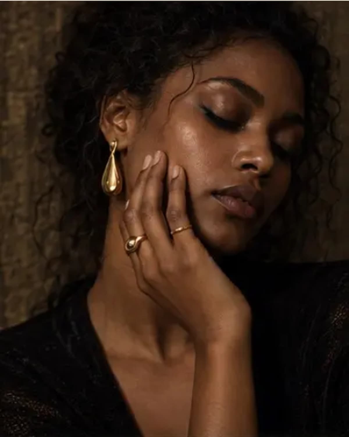 Smiling woman in a black top wearing a teardrop earring, gold watch and pendant necklace, warm daylight